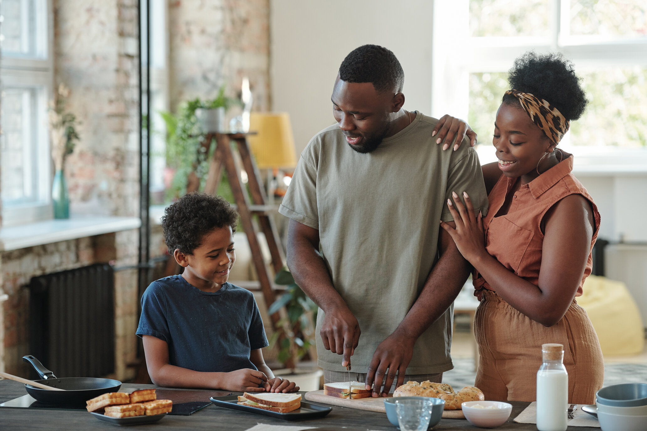 Young African man with his wife and child cutting bread for sandwiches by table in the kitchen for his family